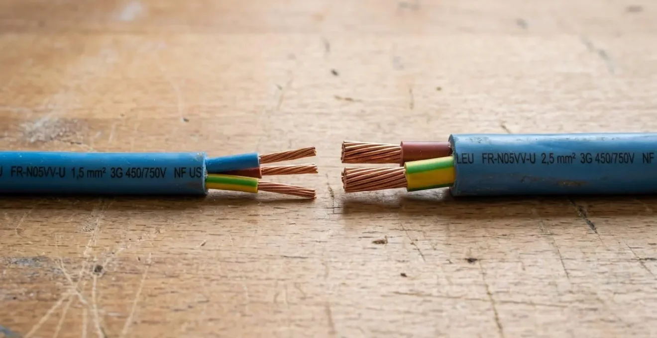 Gros plan sur deux câbles électriques en cuivre de sections différentes (1,5 mm² et 2,5 mm²) placés côte à côte sur un établi, avec différence de diamètre visible et marquages techniques